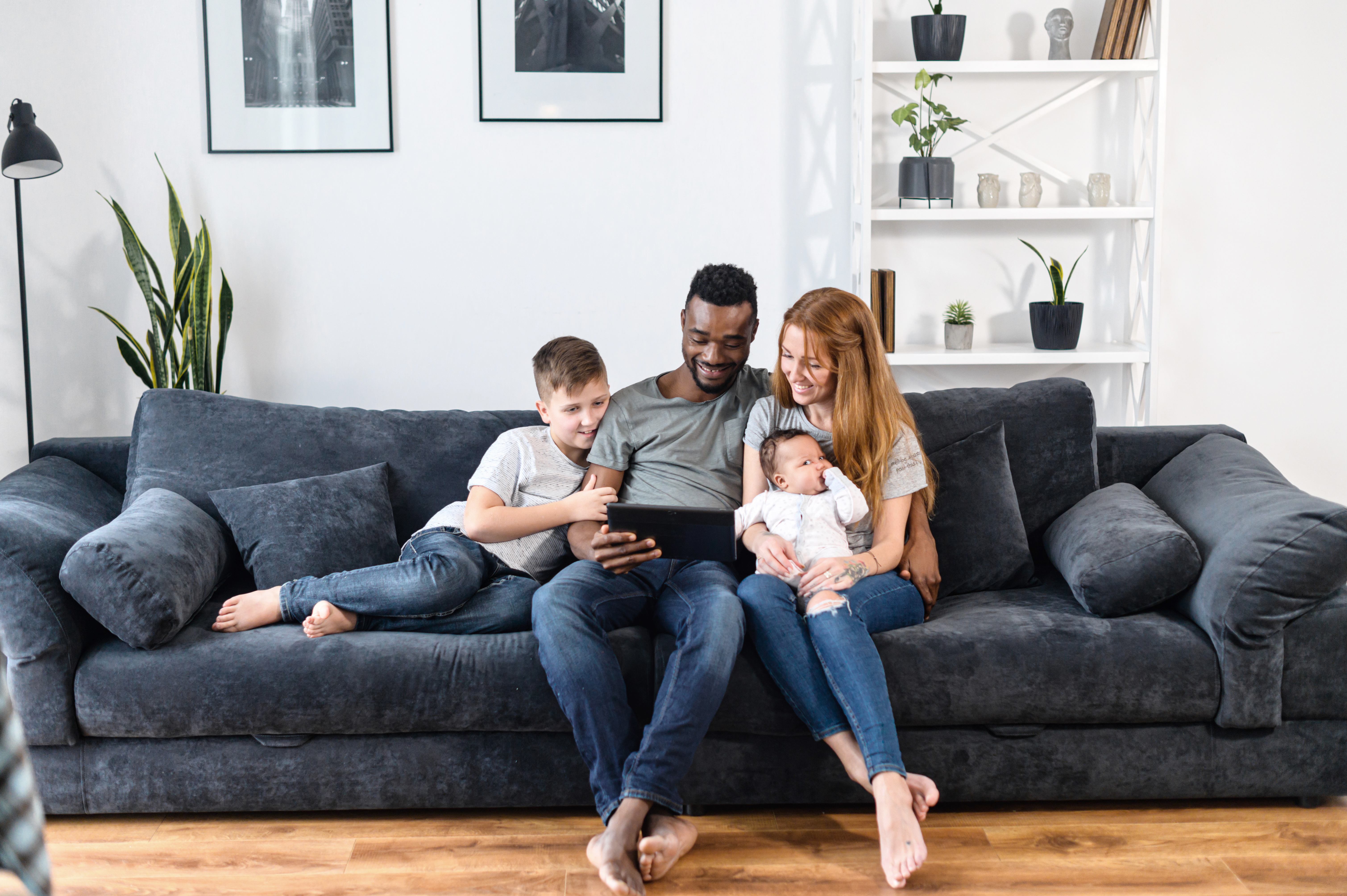 A happy family of four sitting together on a cozy grey sofa, engaging with a tablet. The father is holding the tablet, while a young boy sits beside him and a woman holds a baby in her lap. Soft lighting and indoor plants create a warm atmosphere.