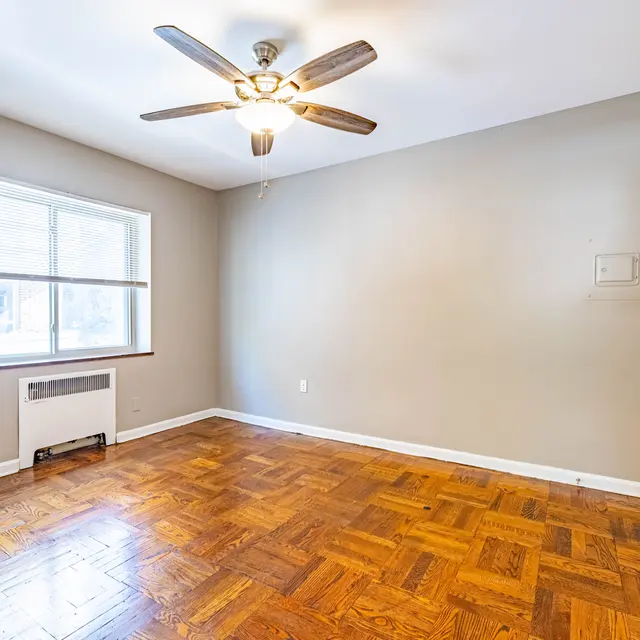 An empty room featuring a wooden floor, a ceiling fan, and a window allowing natural light. There is a wall-mounted heater near the window and a door to the right.