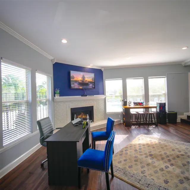 A bright and spacious living area featuring a desk with blue chairs, a cozy fireplace, and large windows letting in natural light. A television is mounted on the wall with a scenic view displayed.