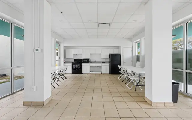 An empty kitchen space with white walls, tiled flooring, and an adjacent dining area. The kitchen features white cabinetry, a stove, and a black refrigerator. Several white tables with chairs are arranged neatly in the dining area.