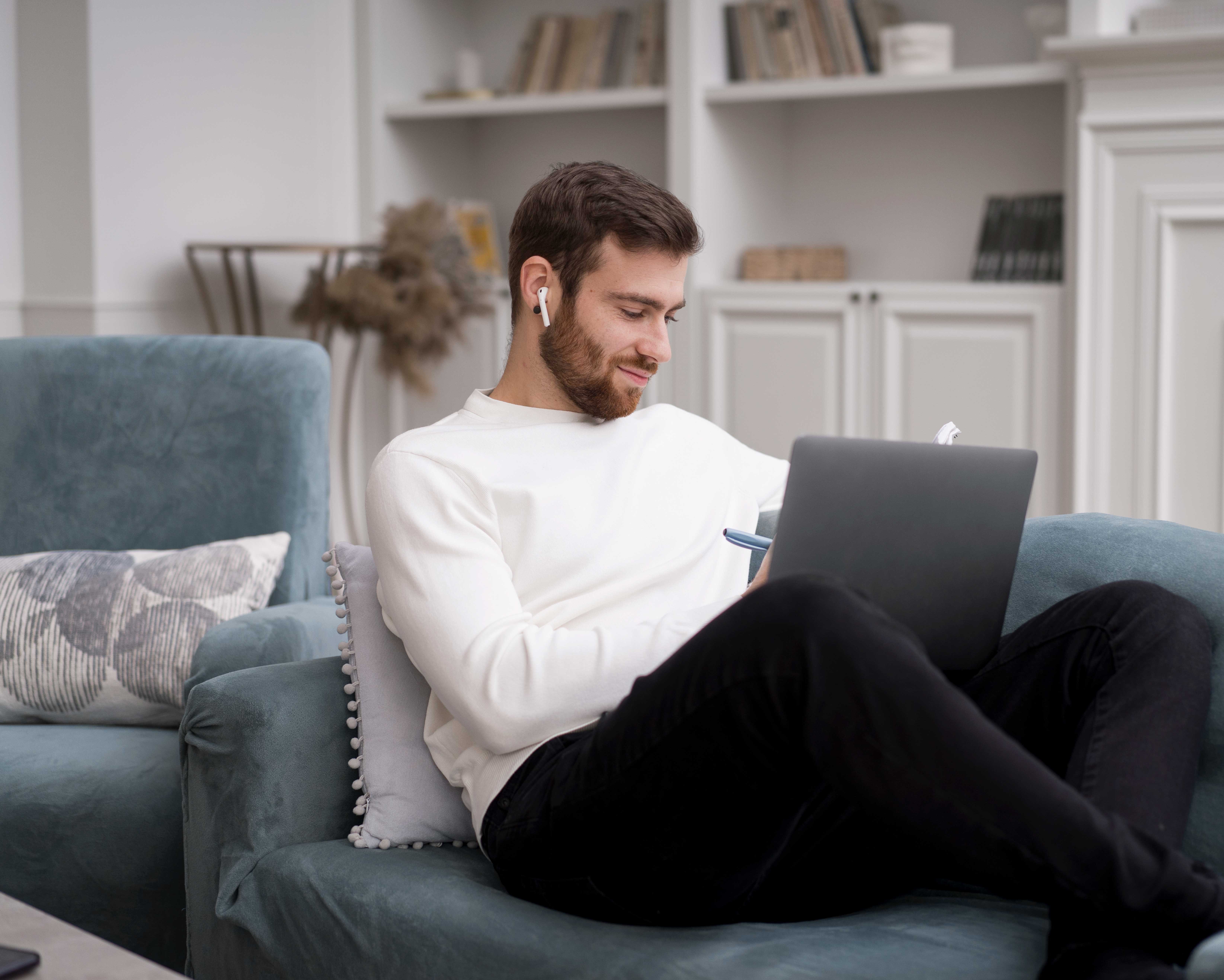 A young man sitting comfortably on a couch, using a laptop. He is wearing a white sweater and wireless earbuds, looking down at his computer with a focused expression. The room is cozy, with bookshelves in the background.