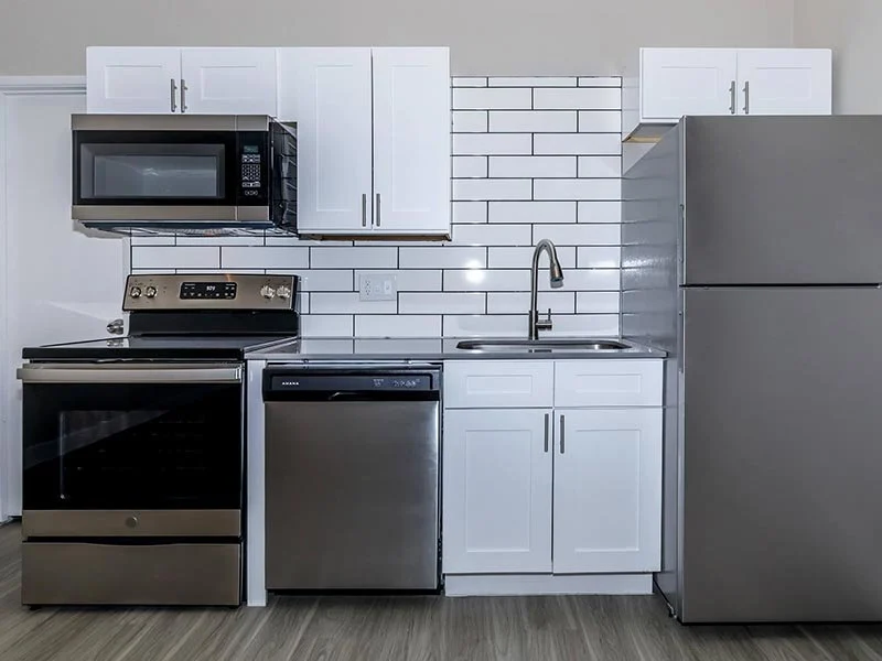 A modern kitchen layout featuring a stainless steel refrigerator, an oven, a dishwasher, a microwave, and white cabinets with a tiled backsplash.