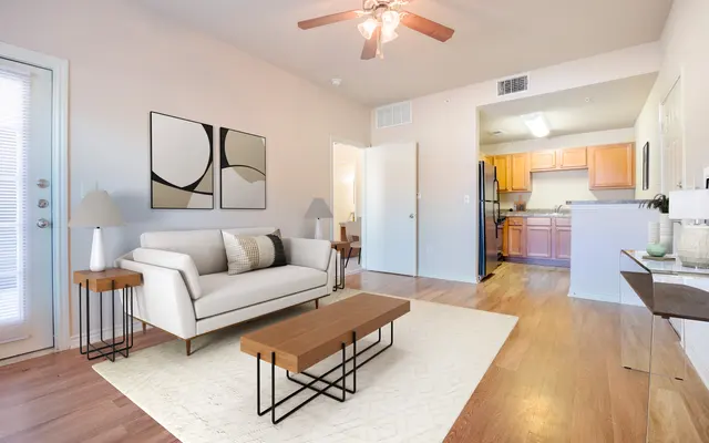 A modern living room with a light-colored sofa, wooden coffee table, and stylish lamps on side tables. The decor includes abstract wall art and a view into a kitchen area in the background.