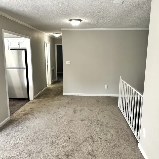 Spacious interior room featuring light gray walls and carpeted flooring, with a view of a staircase railing on the right and an entrance to a kitchen on the left.