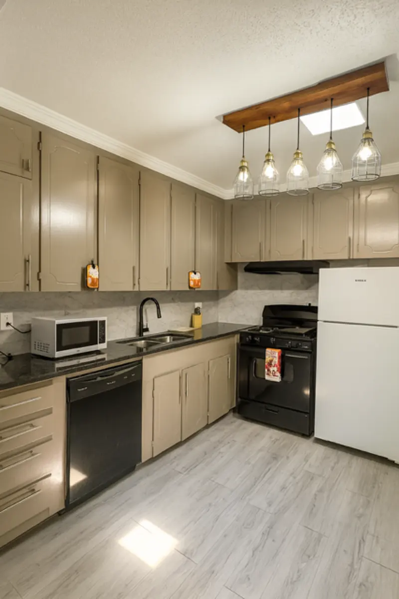 A modern kitchen featuring beige cabinets, black appliances, and a light wood ceiling fixture with hanging bulbs. The countertop is dark and there is a skylight letting in natural light. A microwave and dishwasher are also visible.