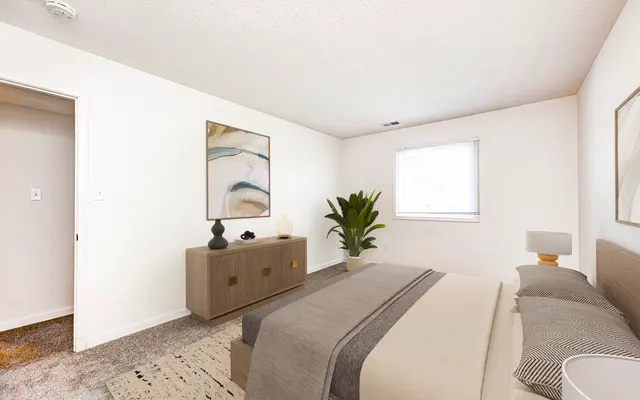 Modern Bedroom Interior A modern bedroom featuring a bed with gray bedding, a bedside lamp, and a dresser with decorative items. A plant and abstract art piece are present, with a window allowing natural light.