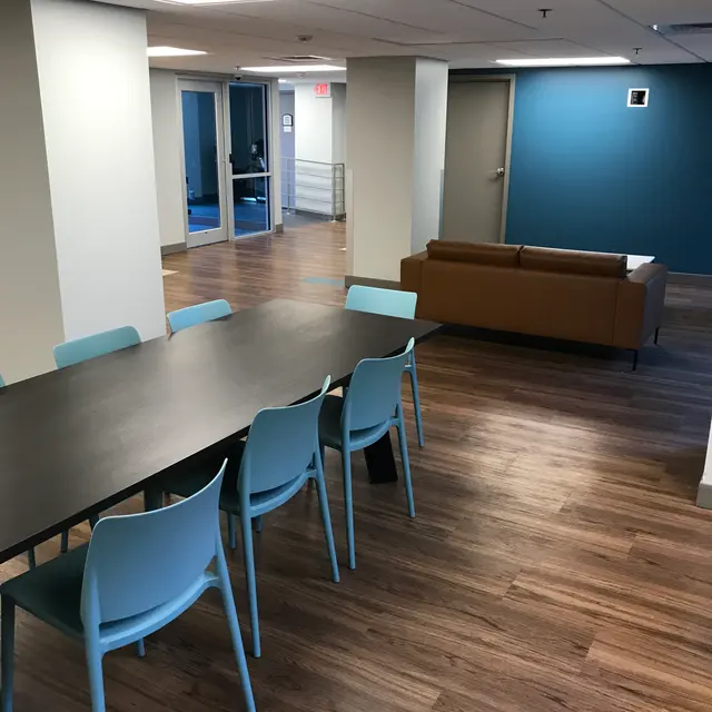 A spacious modern office area with a large table surrounded by light blue chairs. A brown sofa is positioned nearby, and glass doors lead to another area. The floor is wooden, and one wall is painted a vibrant blue.