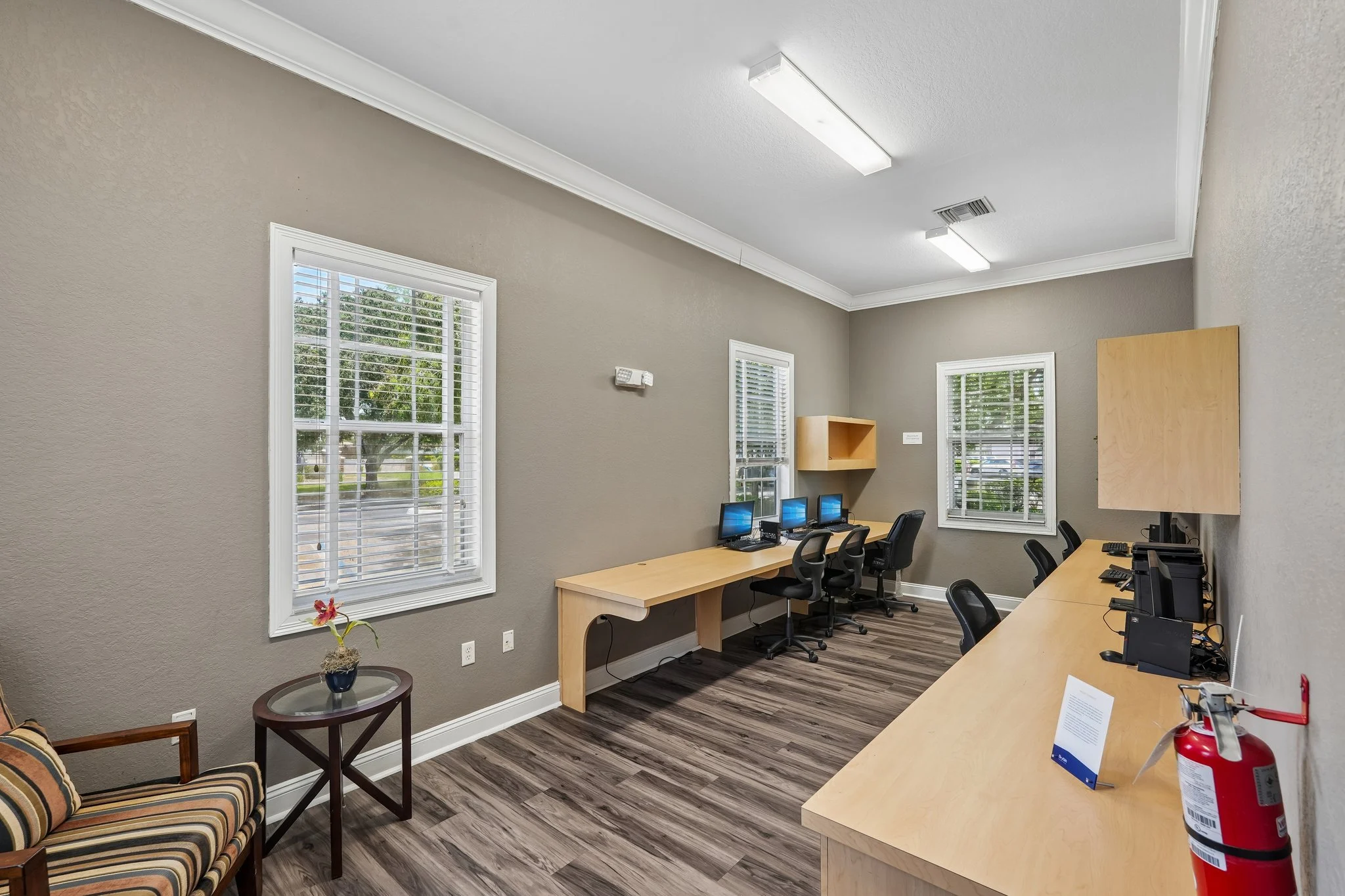 A small office space featuring several desks with computers, a chair, and natural light coming from windows. The walls are a muted color with wood flooring.