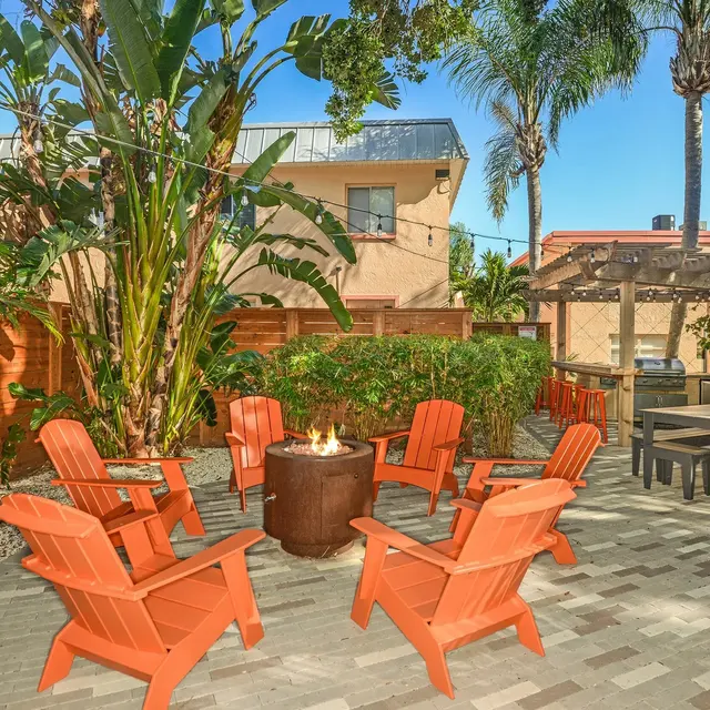 An outdoor seating area featuring orange Adirondack chairs arranged around a fire pit, surrounded by greenery and palm trees.