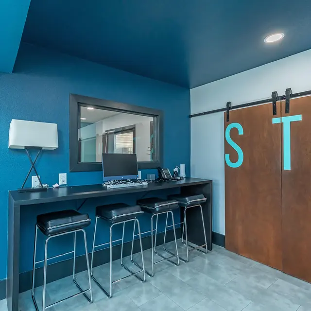 A modern reception area featuring a blue wall, a wooden sliding door with the letters 'STA', and a counter with four stools. A computer setup is visible on the counter, alongside a potted plant in the corner.