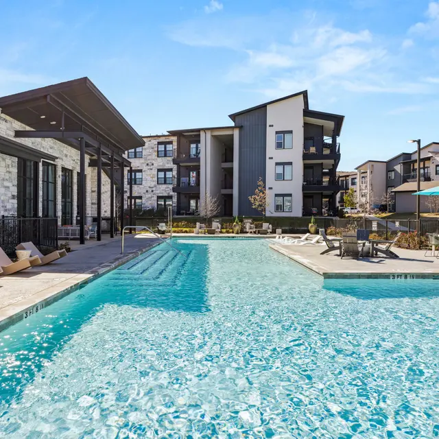 A panoramic view of a swimming pool at an apartment complex, featuring lounge chairs along the poolside and modern buildings in the background.