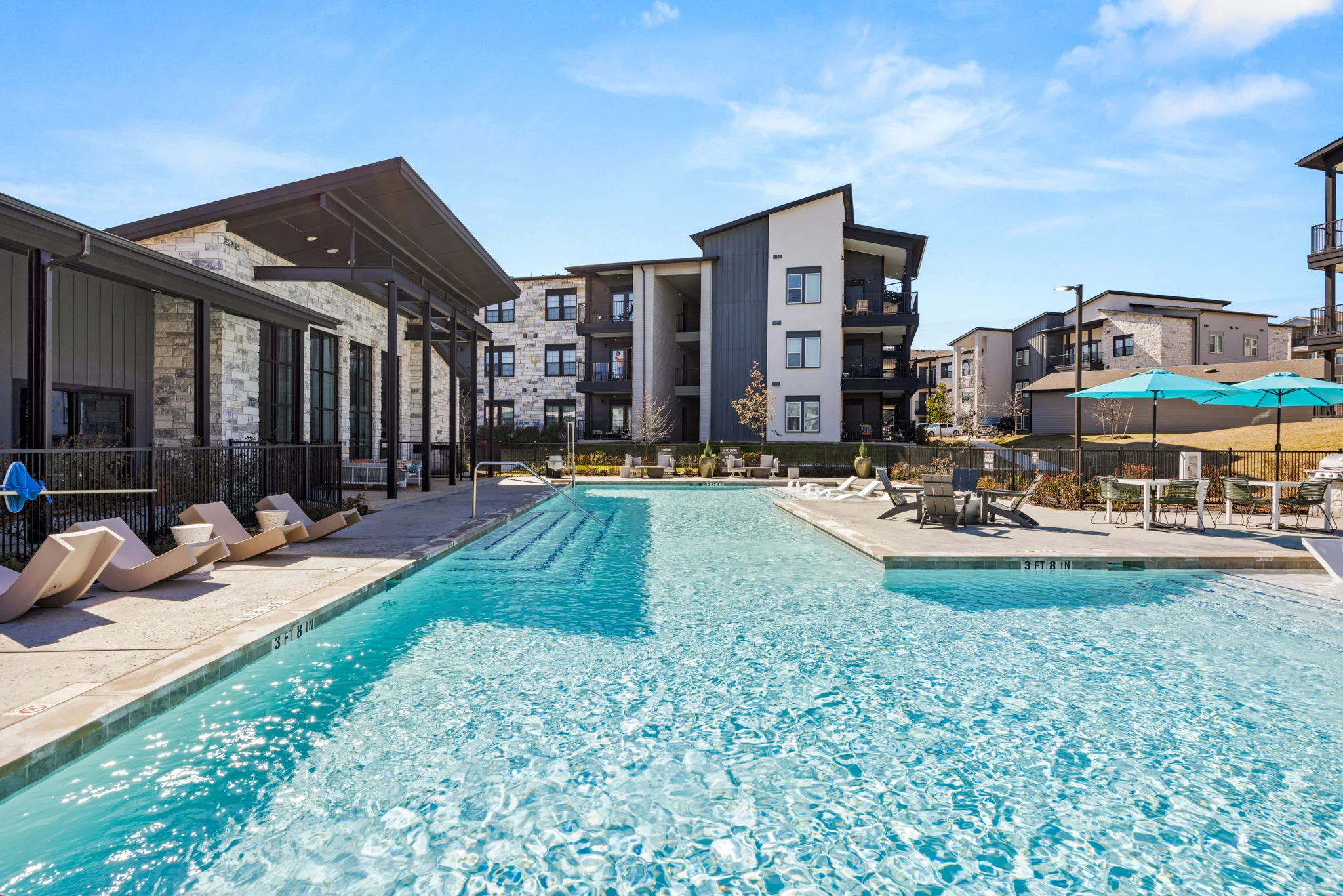 A panoramic view of a swimming pool at an apartment complex, featuring lounge chairs along the poolside and modern buildings in the background.