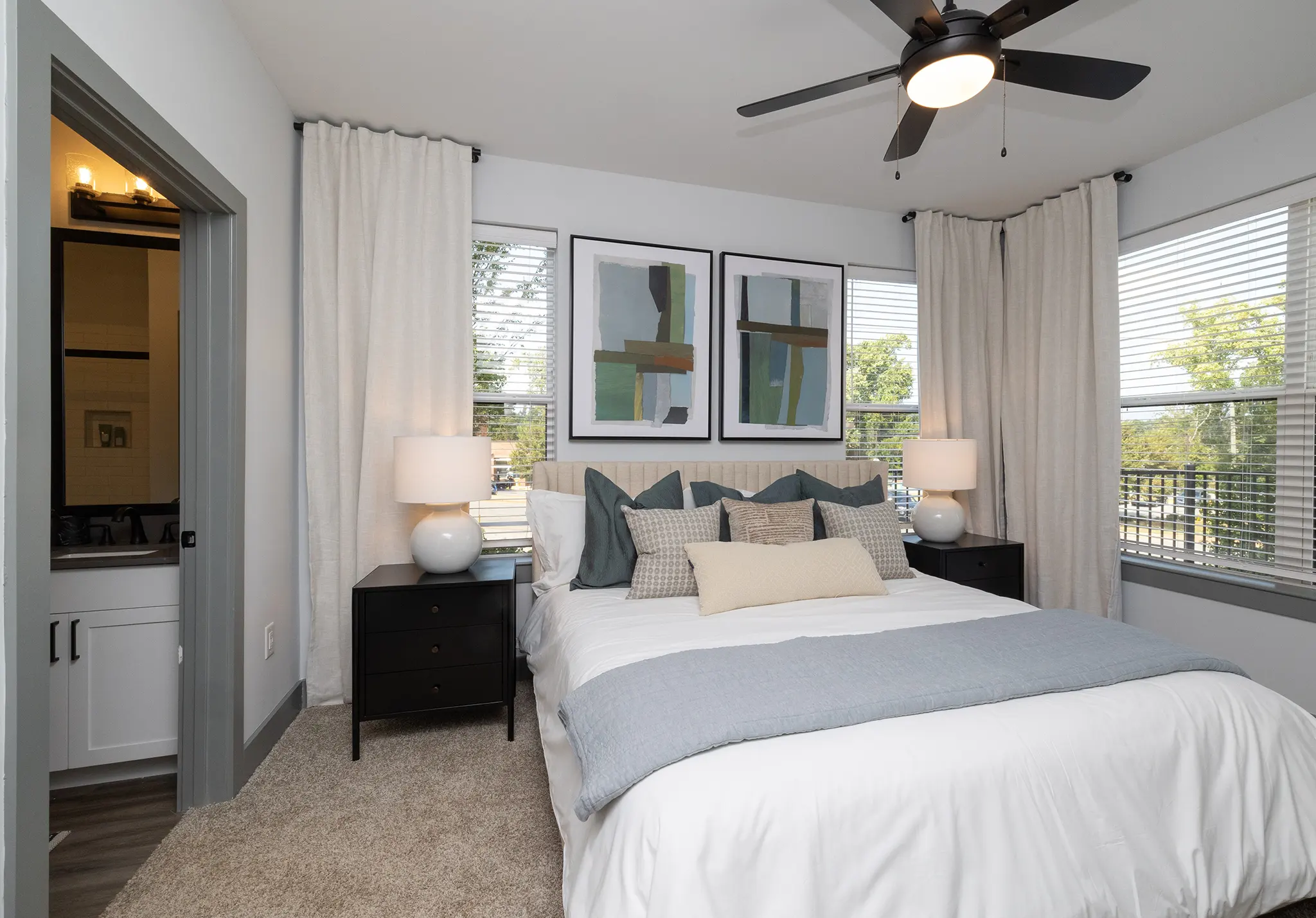 A modern bedroom featuring a comfortable bed with decorative pillows, two nightstands with lamps, and large windows with white curtains overlooking greenery.