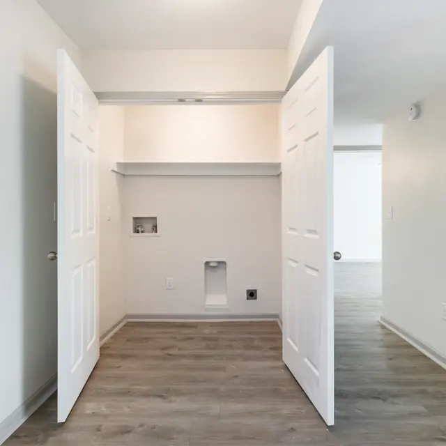 An empty room with open double doors revealing a closet space and a small opening in the wall, featuring light-colored walls and wooden flooring.