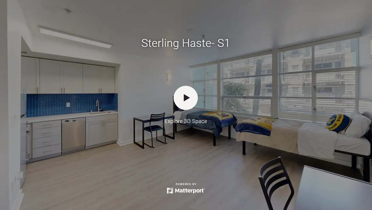 Interior view of a student accommodation room featuring two single beds, a kitchenette with cabinetry, and large windows letting in natural light.