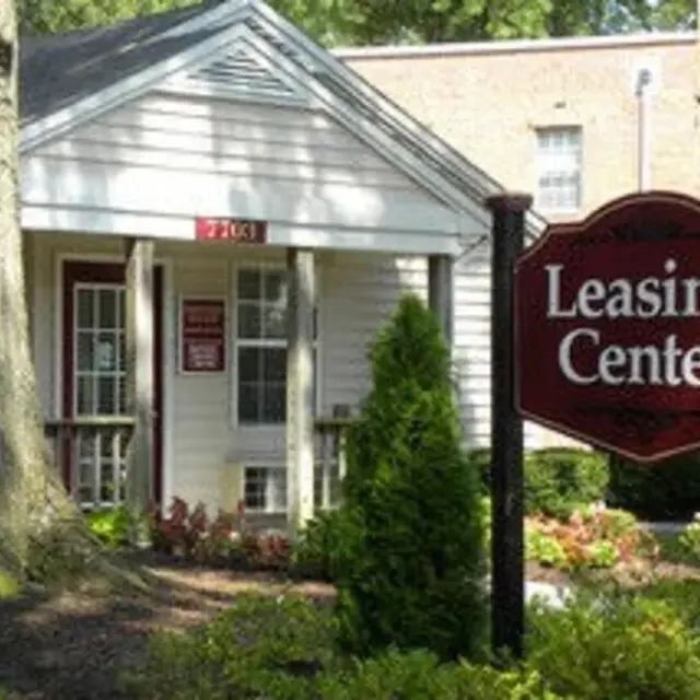 A leasing center building with a sign in front that says 'Leasing Center'. The building is surrounded by greenery and flowers, and there is a tree nearby.