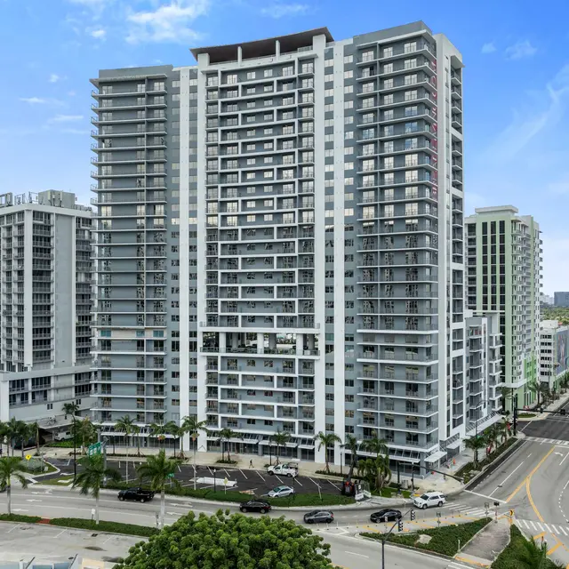 A modern high-rise building with a reflective facade and multiple balconies, surrounded by palm trees and streets.