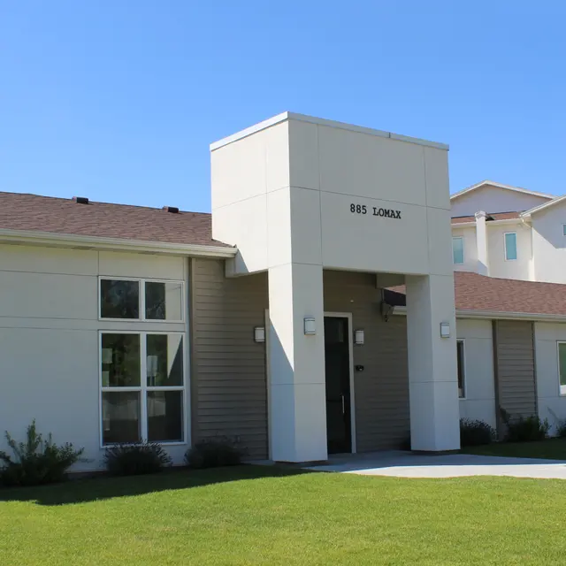 A modern building with a light-colored exterior, featuring a prominent entrance labeled '811 Loma.' The structure has large windows and a sloped roof, surrounded by green grass and landscaping.