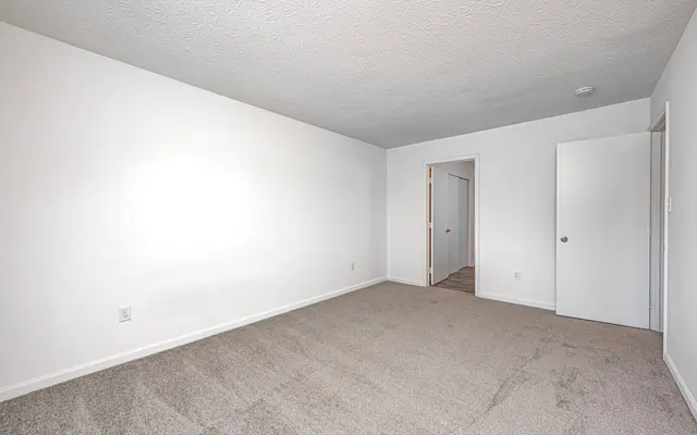 A spacious and empty room with light colored walls and carpeted floor. There is a doorway visible on the left and a closed door on the right, leading to another area.