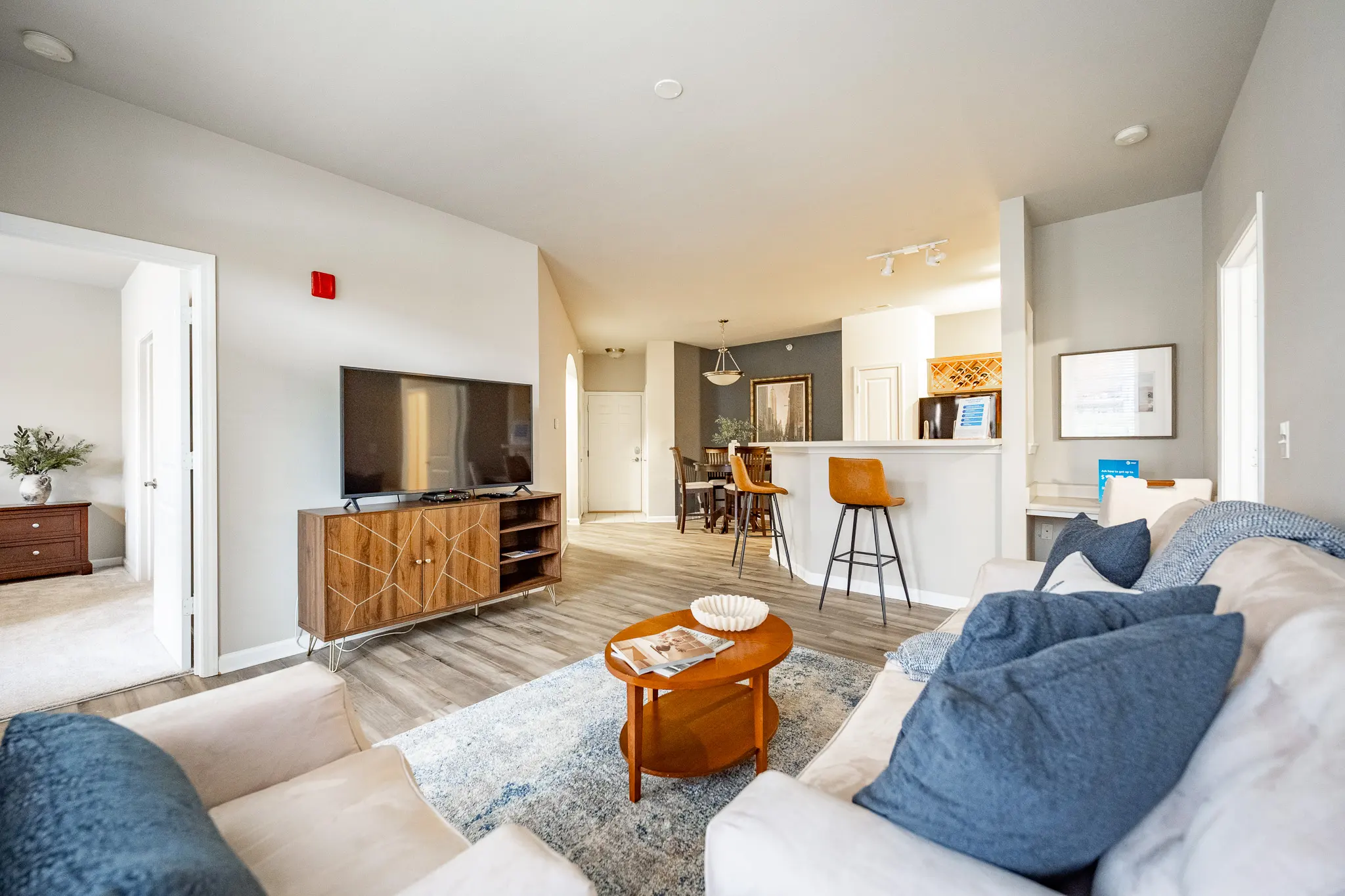 A bright and modern living room featuring a comfortable sofa, a coffee table, and a television unit. The space is open and airy, with a view into a kitchen area that includes bar seating.