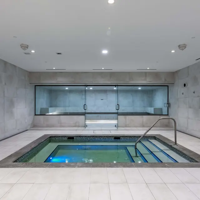 An indoor spa area featuring a rectangular hot tub with a metallic handrail. The walls are smooth and light grey, with large tiles on the floor. In the background, there are glass doors and a clock on the wall, contributing to a serene atmosphere.