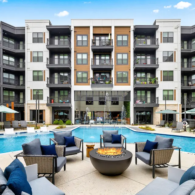 A modern apartment complex featuring a large swimming pool surrounded by lounge chairs and a fire pit. The complex includes multiple balconies overlooking the pool area, with umbrellas and cabanas for shade.