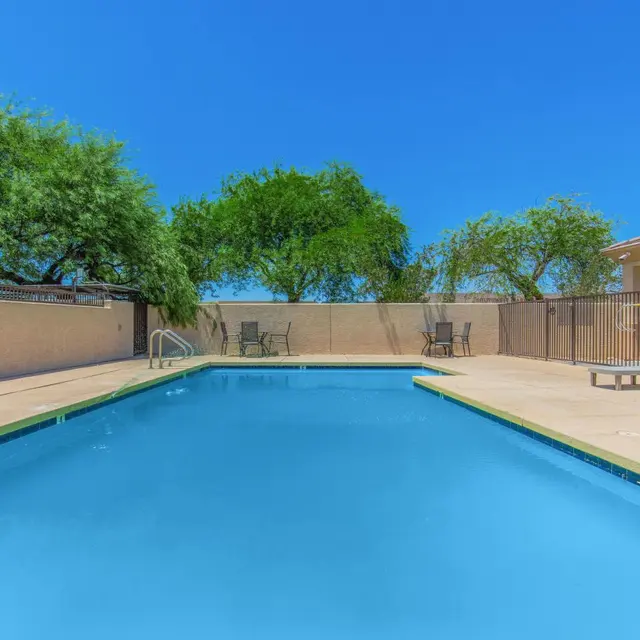Clear Blue Swimming Pool Area A clear blue swimming pool surrounded by sun loungers and fencing, with trees and a building in the background under a bright blue sky.