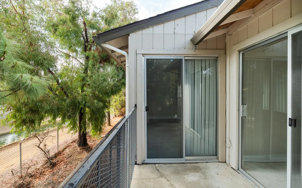 A spacious balcony with a sliding glass door, featuring a view of green trees and a fenced area below. The flooring is concrete and the surrounding area is serene, suggesting a tranquil outdoor space.