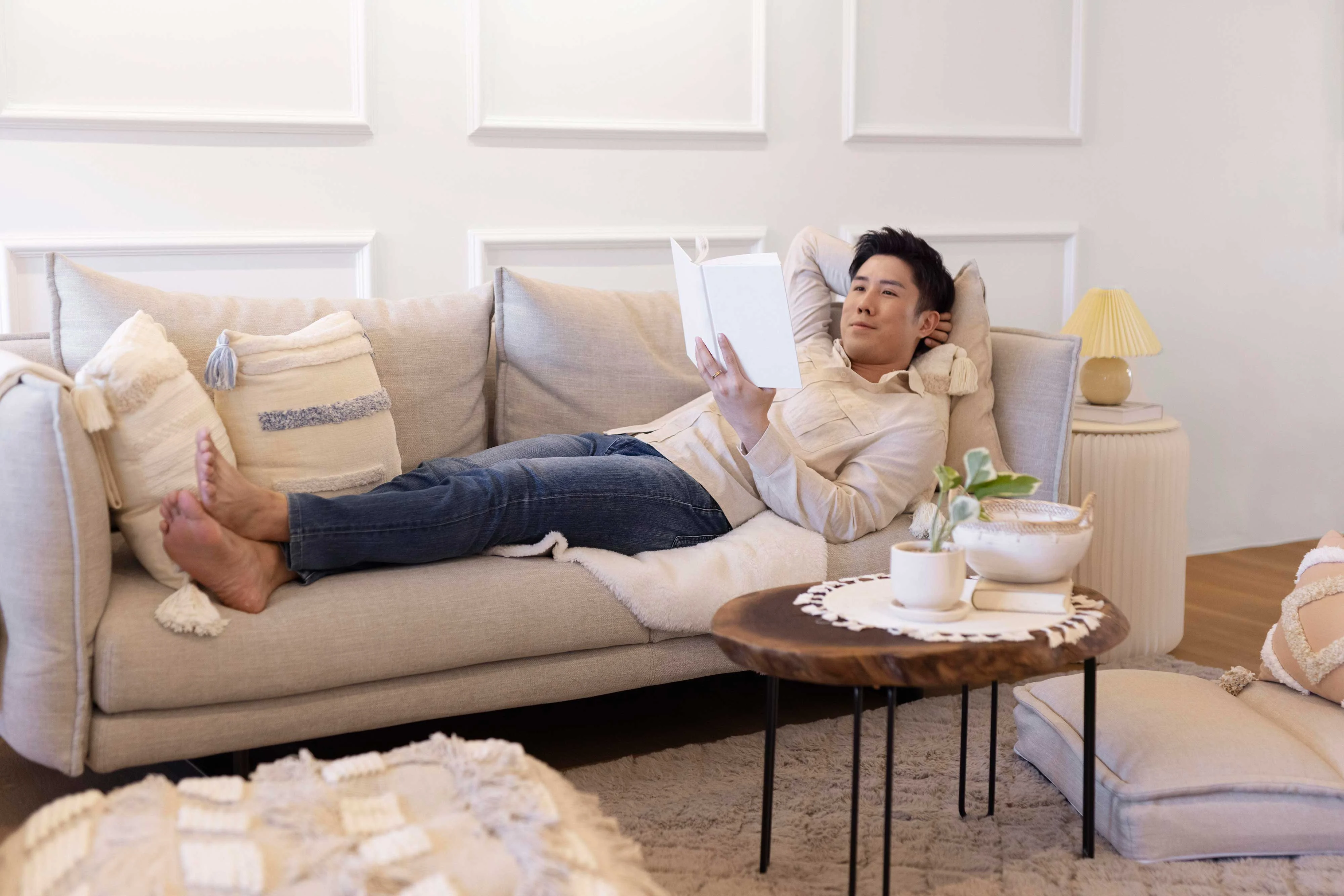 A man lounging on a light gray sofa, reading a book. He is barefoot and relaxed, wearing a light-colored shirt and blue jeans. The room is bright and decorated with cushions and a small coffee table holding a plant and a cup.