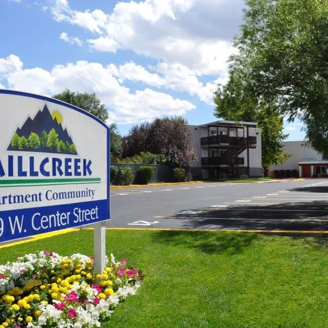 A sign for Millcreek Apartment Community with flowers in the foreground and an apartment building in the background on a sunny day.