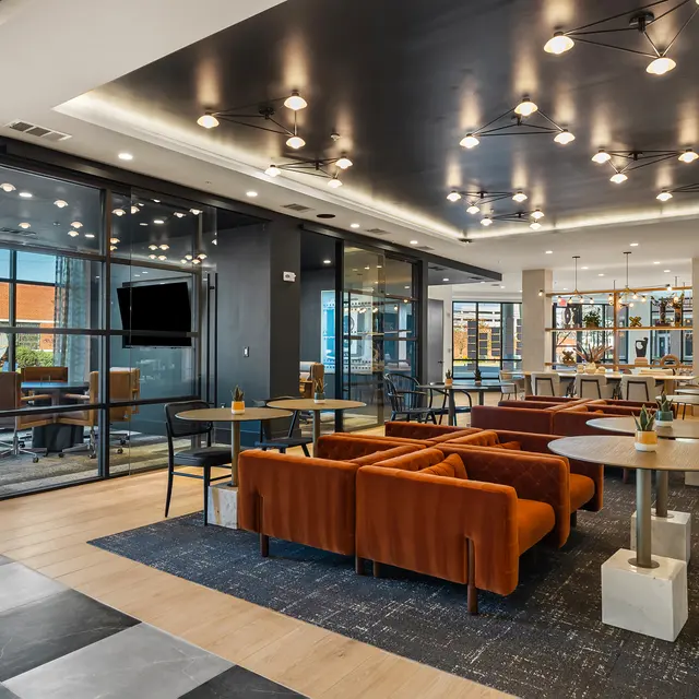 A stylish co-working space featuring orange sofas, wooden tables, and glass meeting rooms with modern lighting.