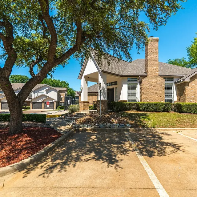 A charming brick house with a sloped roof, surrounded by trees and landscaped bushes. The driveway is paved and has a few parking spaces.