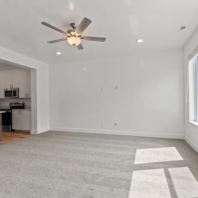 A spacious living room with carpet flooring and large windows allowing natural light. There is a ceiling fan, and the kitchen area is partially visible with modern appliances and cabinetry.