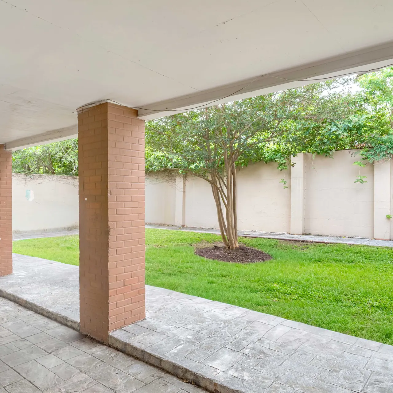 A view of an outdoor space featuring a concrete floor and two pillars supporting the roof. On the other side of the pillars, there is a lush green lawn with a small tree, enclosed by a beige wall. The area is well-lit, suggesting it is daytime.