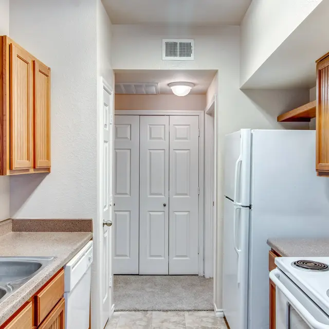 Modern Kitchen Overview A view of a modern kitchen featuring wooden cabinets, a double sink, a white stove, and a refrigerator. The kitchen is well-lit and transitions into a hallway with closed doors.