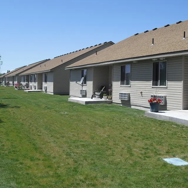 A row of beige houses with brown roofs on a sunny day, surrounded by green grass. Each house has a small porch with chairs and potted plants. Trees are visible in the background.