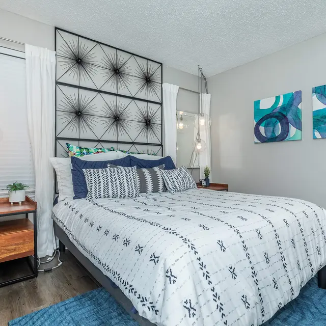 A modern bedroom featuring a bed with a patterned white comforter, decorative pillows, and a geometric wall art piece. The room has two hanging light fixtures, sheer curtains, and two abstract paintings on the wall. A nightstand with decorative items is next to the bed.