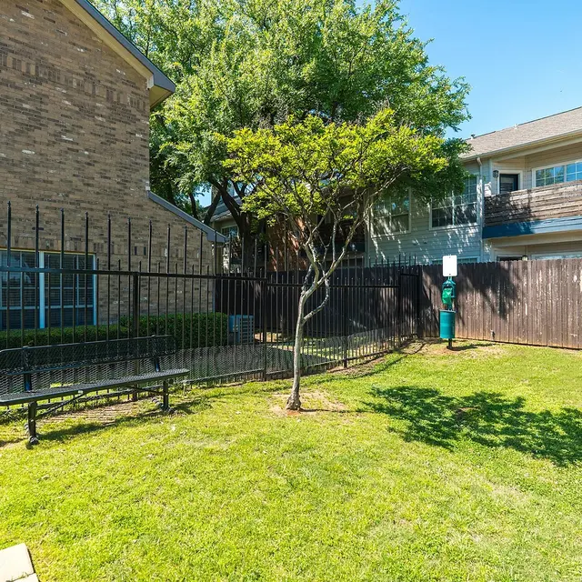 A small, fenced backyard area with a bench, a tree, and green grass, surrounded by buildings in a residential setting.