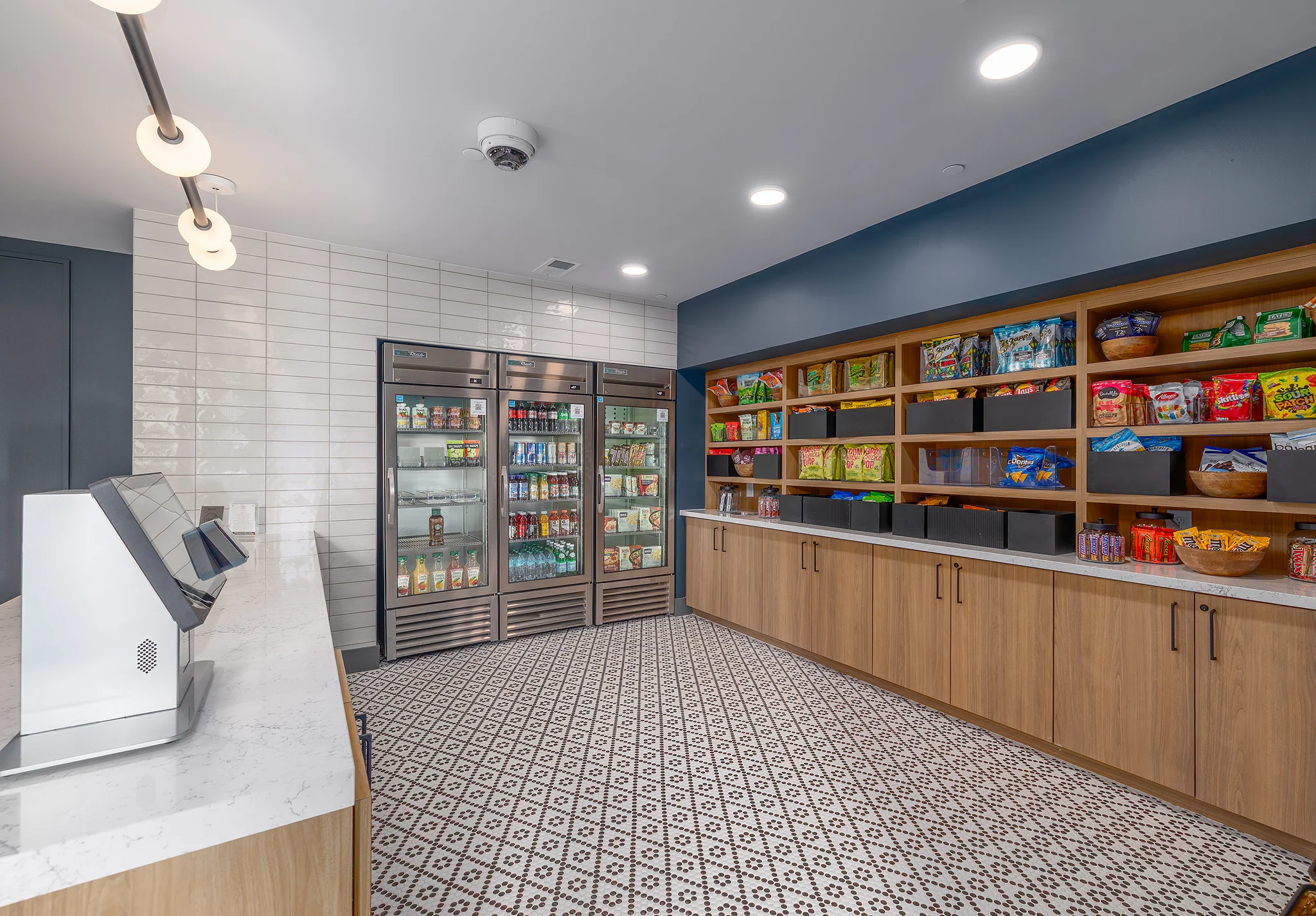 A modern kitchenette space featuring a stylish design with a refrigerator displaying a variety of beverages, wooden shelving stocked with snacks, and an inviting tiled floor.