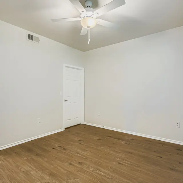 An empty room with light-colored walls and wooden flooring, featuring a ceiling fan. A door is visible on the right side, and a doorway leads to another space.