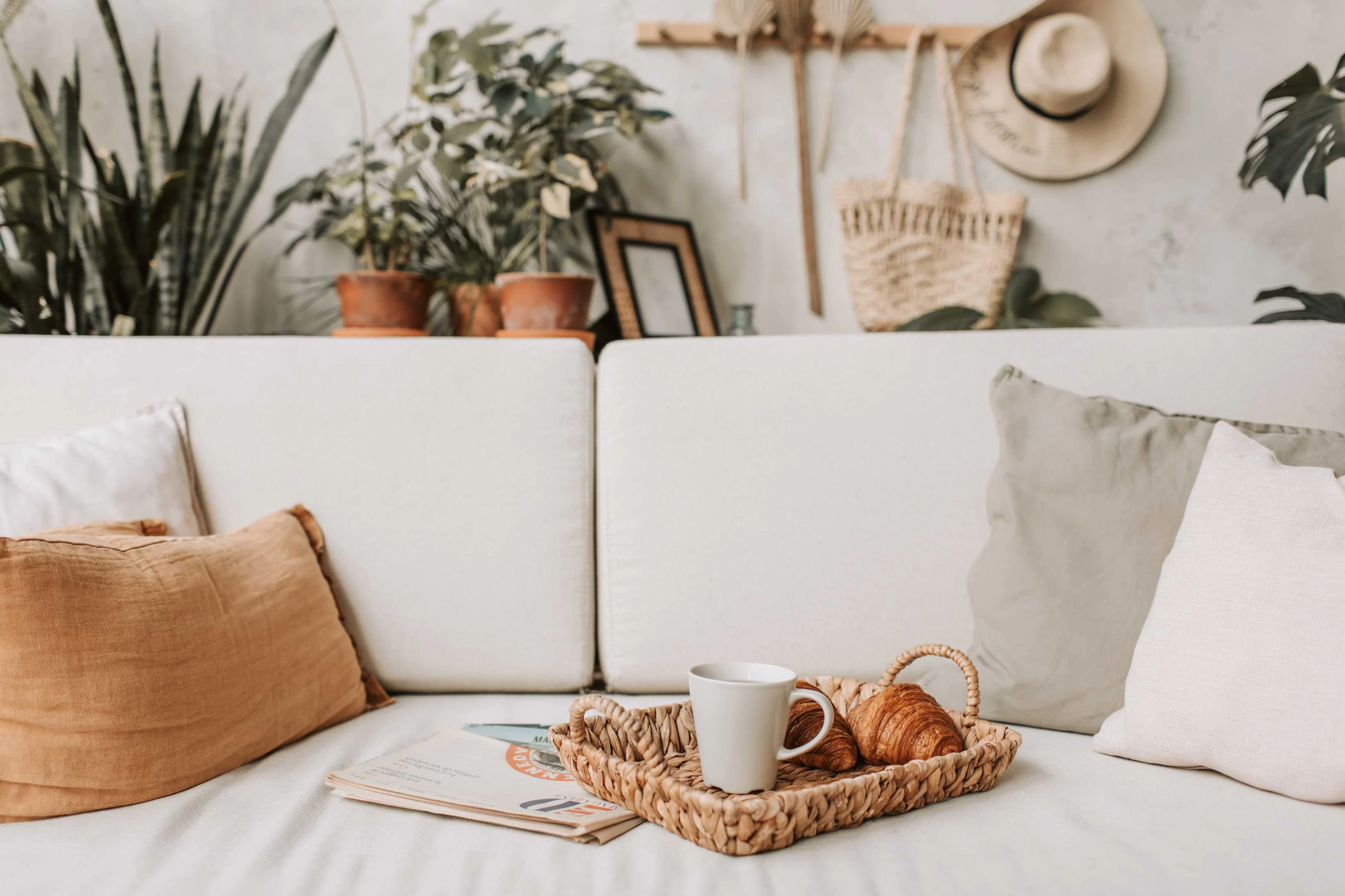 Cozy Living Room Nook A cozy living room scene featuring a white couch adorned with various cushions, a woven tray with a cup and croissants, and greenery in the background.