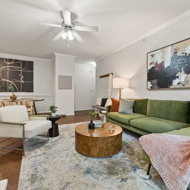 A cozy living room with a green sofa, white chair, round wooden coffee table, and a decorative rug. A ceiling fan and a lamp provide light, with modern artwork on the wall. A dining table is seen in the background.