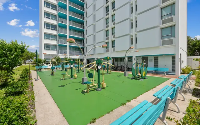 An outdoor exercise area with fitness equipment surrounded by greenery, adjacent to a modern apartment building.