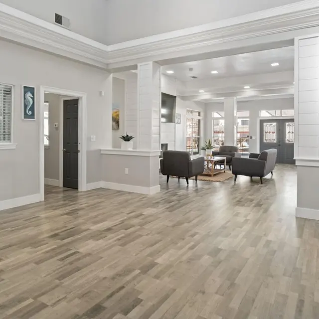 A spacious indoor lobby area with light wood flooring, modern decor, and large windows allowing natural light. A small table with decorative items is on the left, and comfortable seating is arranged in the center.