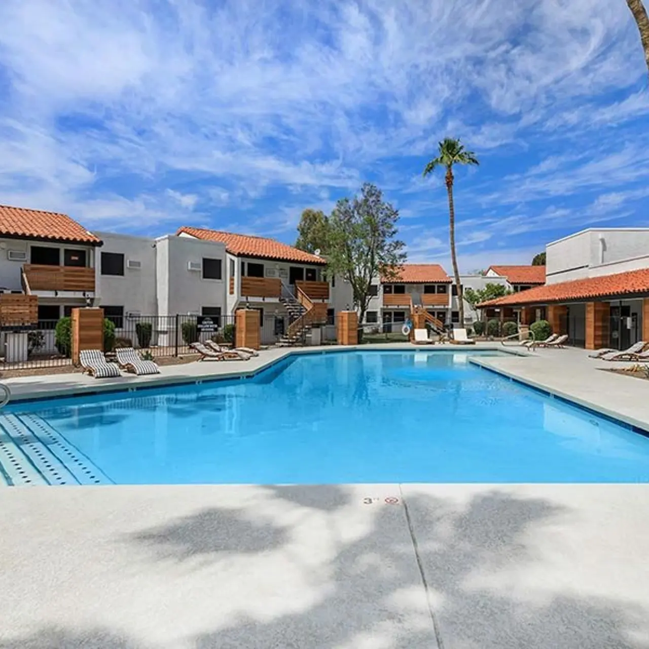 A bright, sunny pool area surrounded by residential buildings in an apartment complex. There are lounge chairs beside the pool and palm trees in the background.