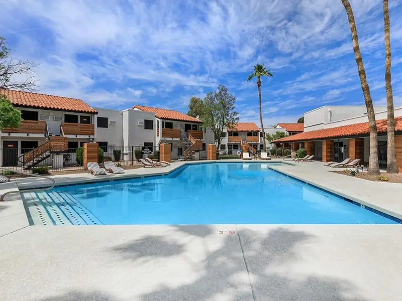 A bright, sunny pool area surrounded by residential buildings in an apartment complex. There are lounge chairs beside the pool and palm trees in the background.