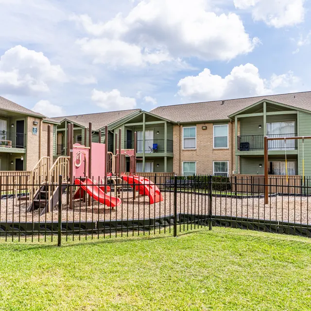 View of a playground surrounded by an apartment complex, featuring slides and a fenced area.