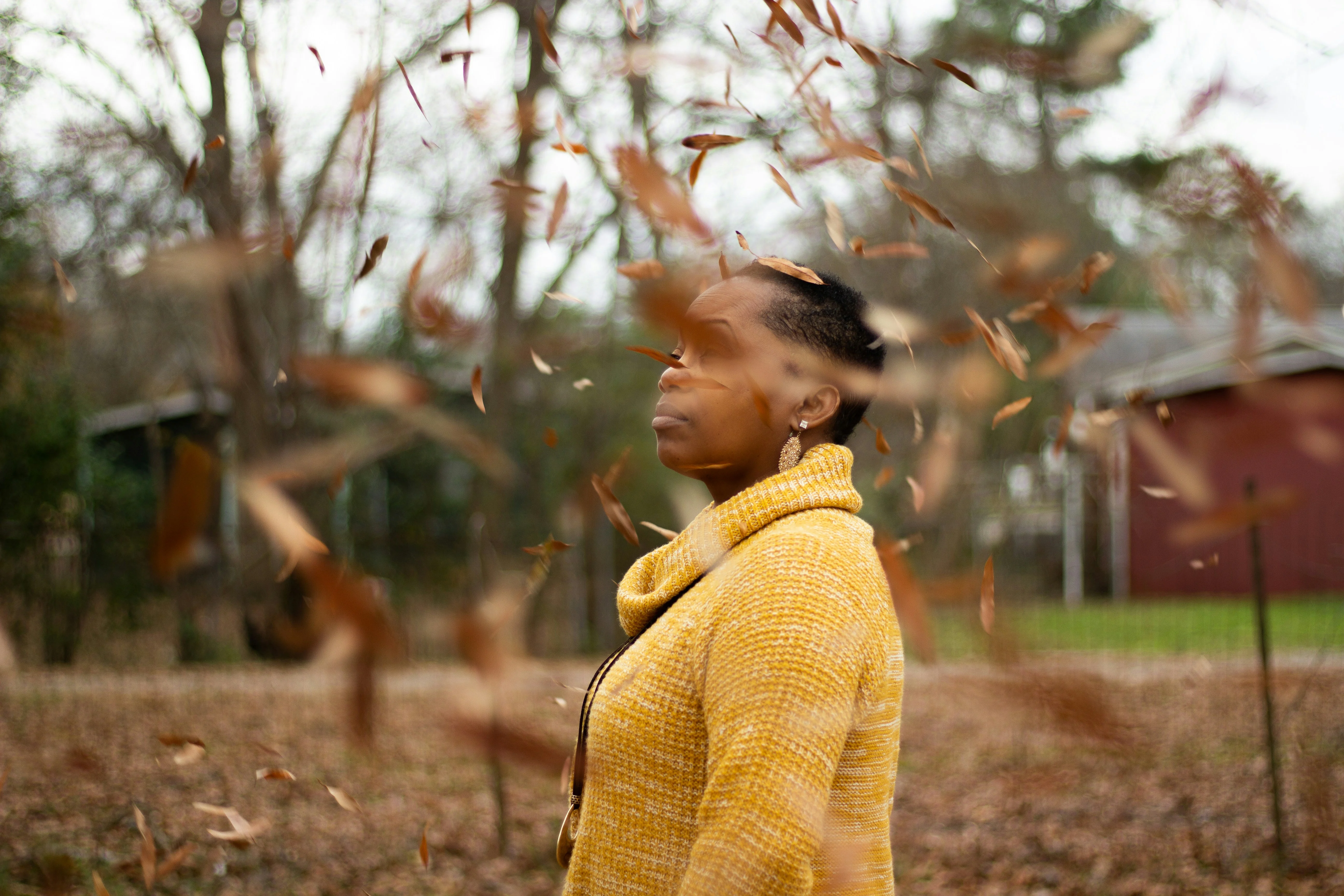 Serene Autumn Moment A woman wearing a yellow sweater stands still with falling leaves around her in a forested area.