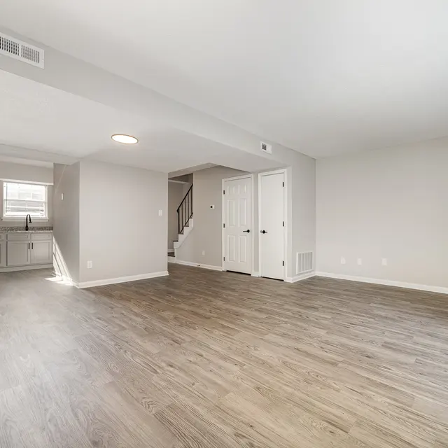 A spacious and modern living room with hardwood flooring, neutral-colored walls, and plenty of natural light. The area features a staircase, a doorway leading to a kitchen, and multiple doors leading to other rooms.