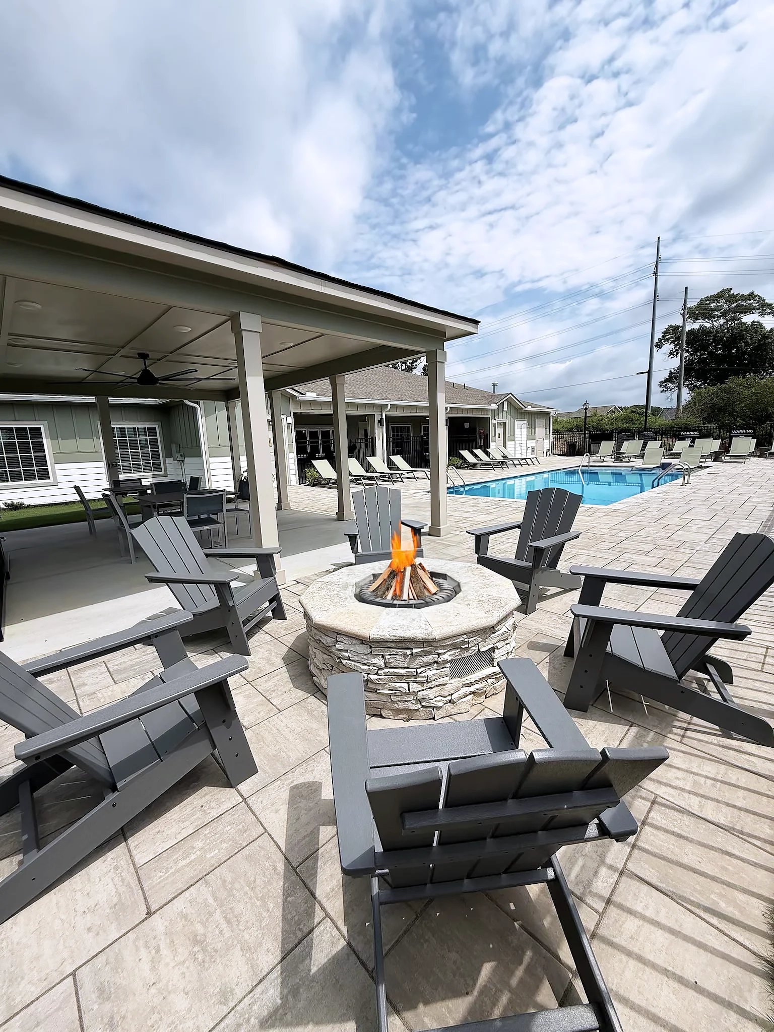 A cozy outdoor area with grey Adirondack chairs surrounding a stone fire pit, next to a swimming pool. The sky is partly cloudy, and there are some trees in the background.